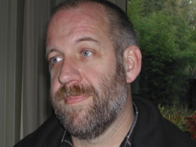 A bearded man with a neutral expression looks slightly to the side in an indoor setting with greenery outside.