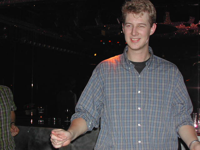 A young man in a plaid shirt smiles while moving his hands, appearing to dance or shake.