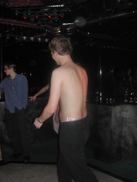 A shirtless man walks in a dimly lit room with money tucked into his pants.