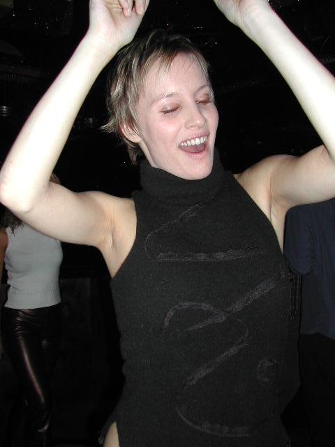 A person with short hair is dancing with arms raised, smiling with eyes closed in a dimly lit setting.