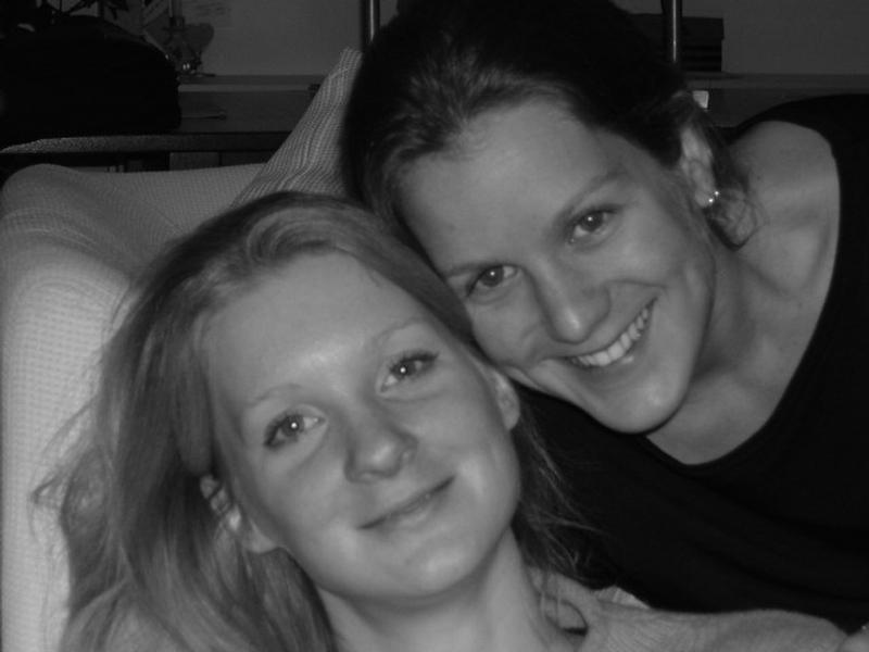 Two smiling women pose closely together, one leaning over the other on a couch.