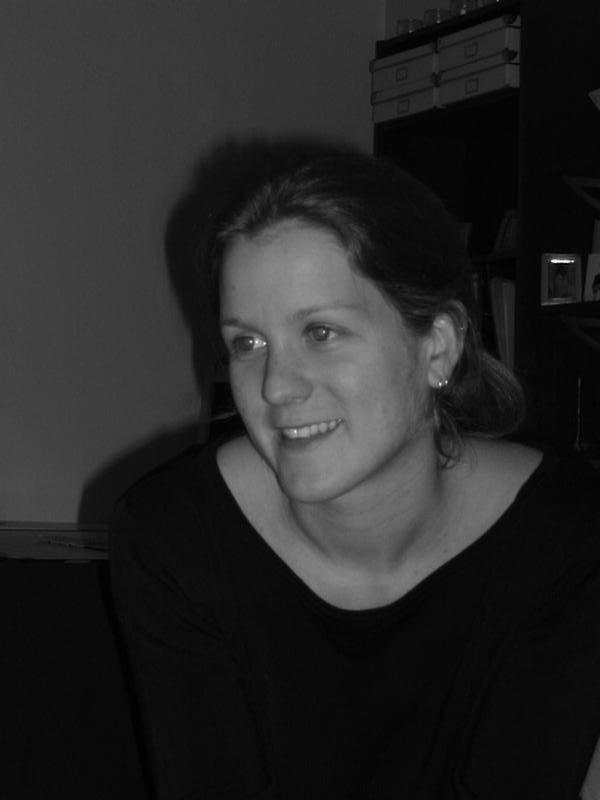 A woman with tied-back hair and a black top smiles while looking slightly to the side in an indoor setting.
