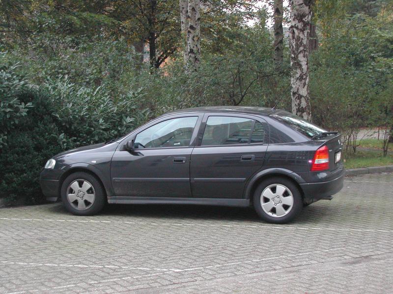 A dark-colored hatchback car is parked on a paved surface near trees and bushes.