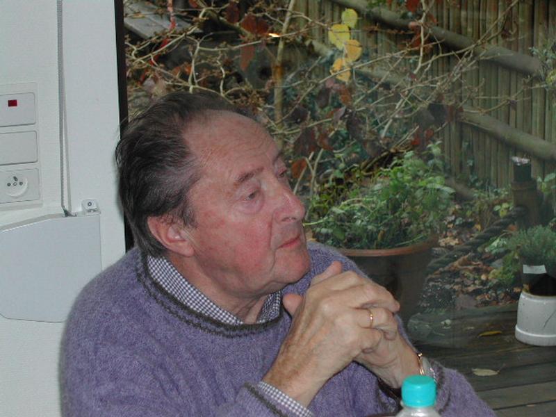 An elderly man in a purple sweater sits indoors, resting his hands together and looking to the side.