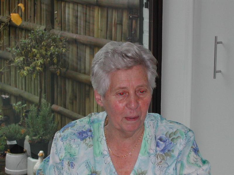 An elderly woman with short gray hair sits indoors, wearing a floral blouse and a gold necklace.