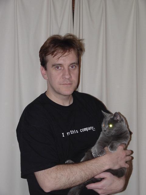 A man in a black t-shirt holds a gray cat while standing in front of a curtain.