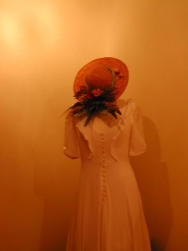 A white wedding dress with buttons down the back, displayed with a straw hat decorated with flowers.