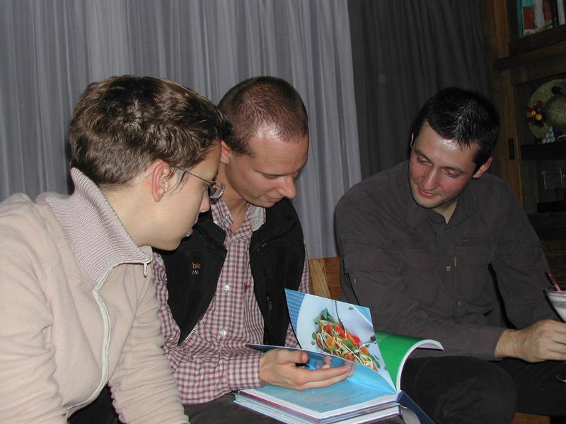 Three people sit together, looking at a book with food images and discussing something with interest.