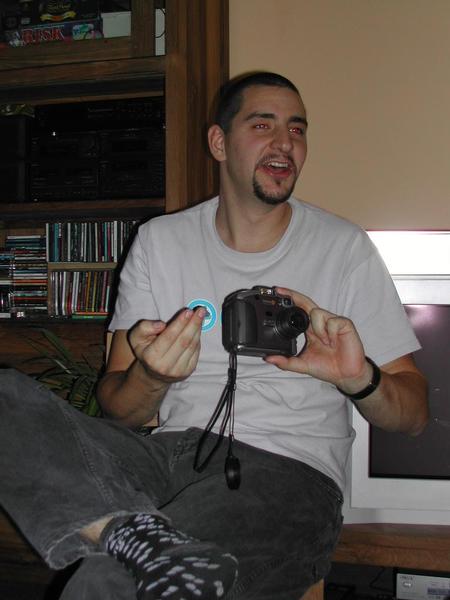 A man in a white shirt laughs while holding a camera and making a playful gesture with his hand.