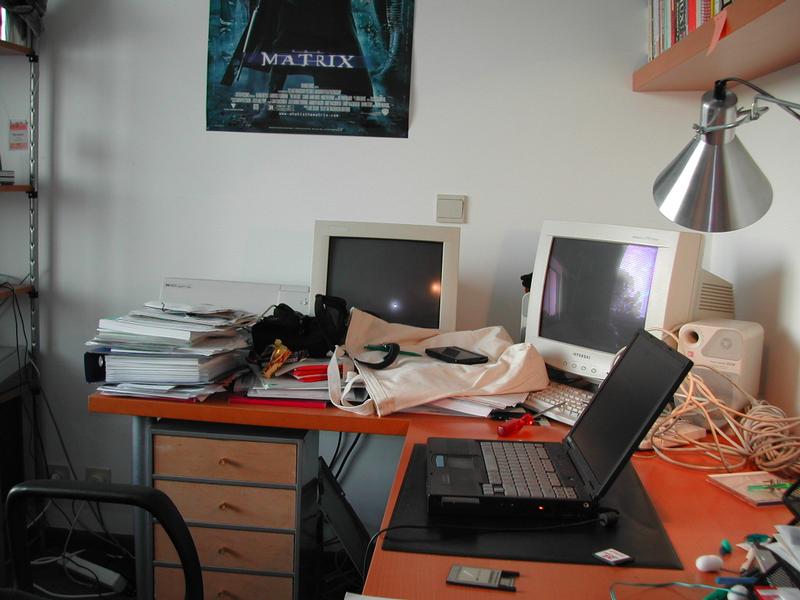 A cluttered desk with an open laptop, two older computer monitors, stacks of papers, and various office supplies.
