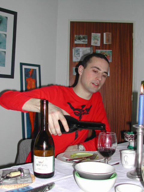 A man in a red shirt pours red wine into a glass at a dining table.