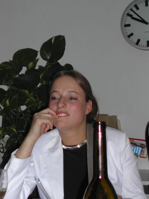 A woman in a white blazer sits at a table, smiling with her hand on her face.