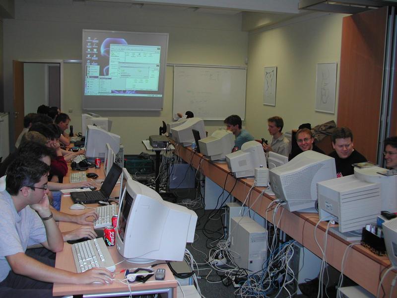 A group of people using desktop computers at a LAN party, with screens, cables, and drinks on the desks.