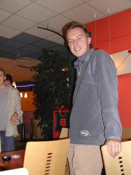 A man in a gray jacket smiles while leaning on a chair in a casual indoor setting.