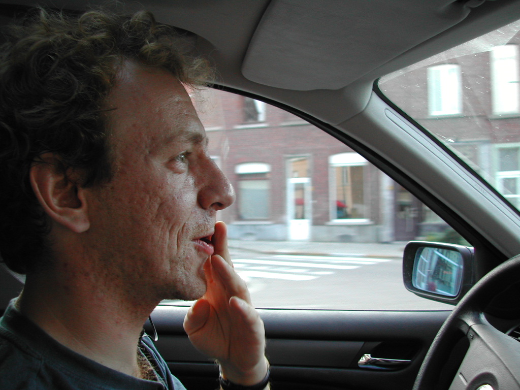 A man is driving a car, touching his face with one hand while looking ahead.