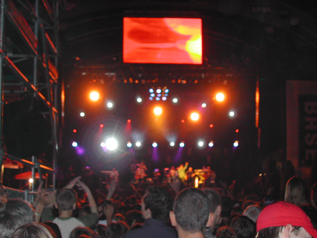 A large crowd watches a live concert at night, with bright stage lights and a big screen above.