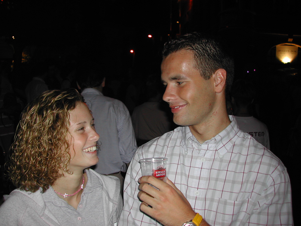 A young man and woman smile at each other while he holds a cup of beer at an outdoor event.