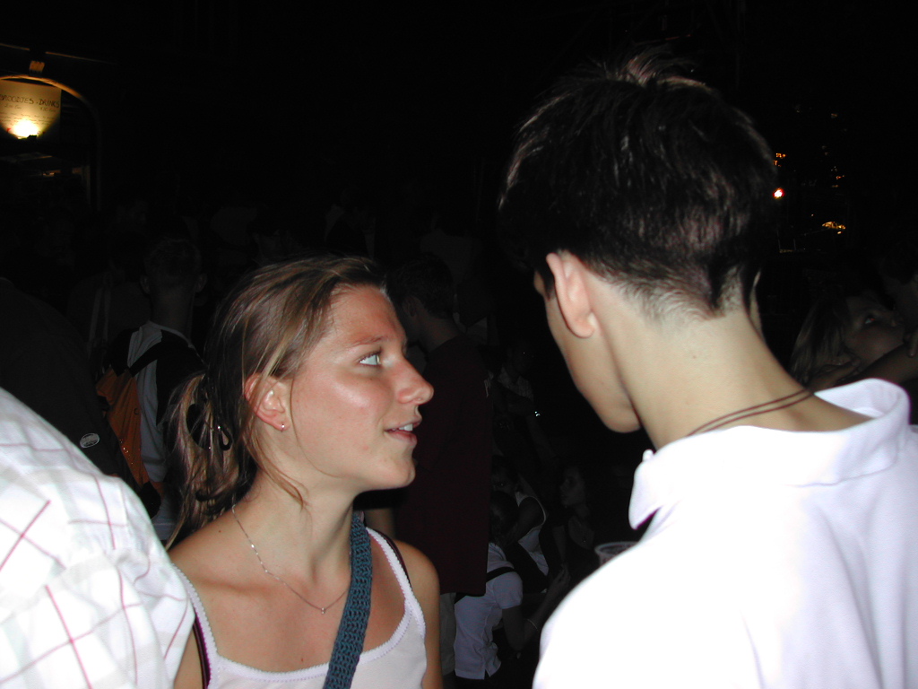 A woman and a man are talking in a crowded, dimly lit outdoor setting.