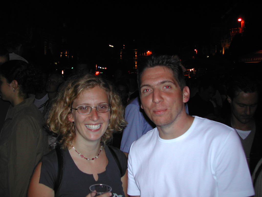 A smiling woman holding a drink and a man in a white shirt stand together at a crowded nighttime event.