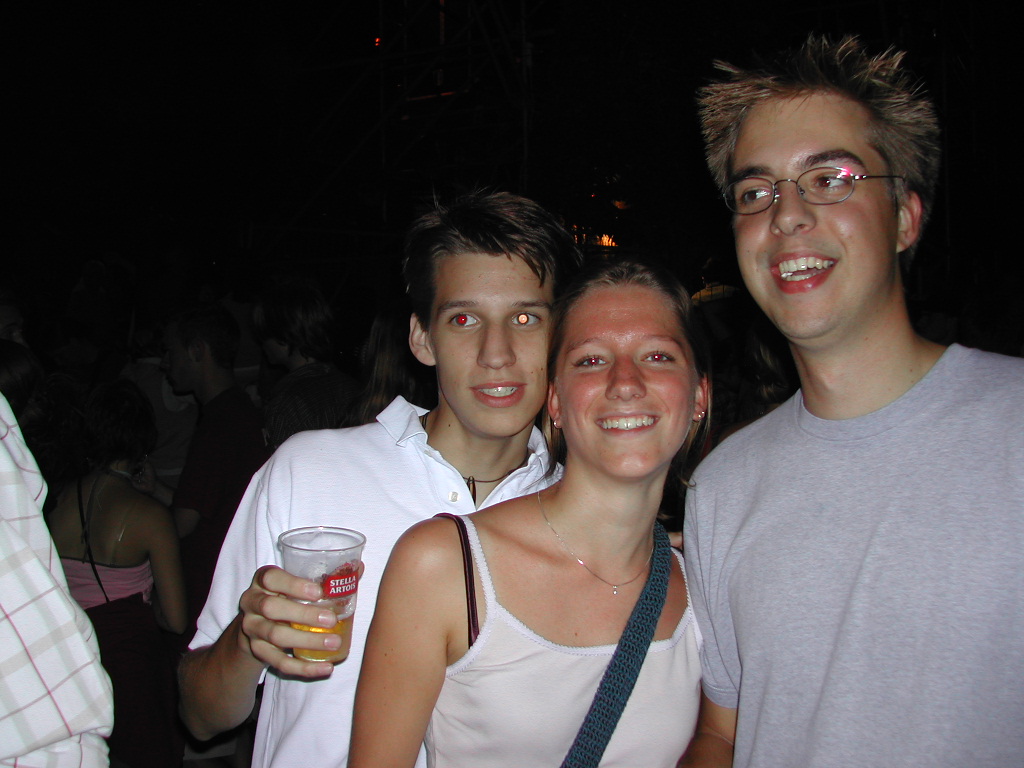Three young adults smile at the camera at a crowded nighttime event, one holding a cup of beer.