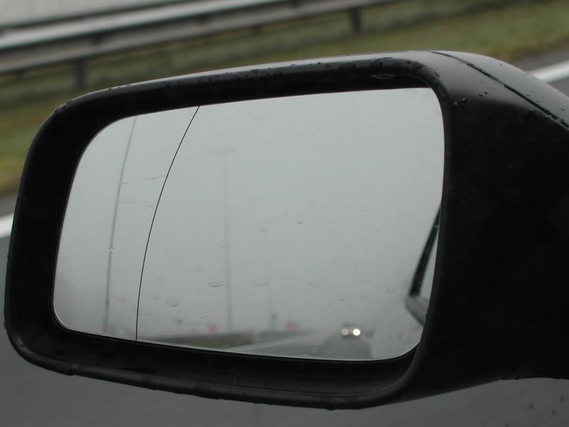 A car's side mirror with raindrops and a visible crack reflects a foggy road with a distant vehicle.
