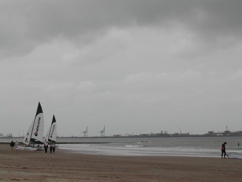 People stand near two sailboats on a cloudy beach, while another person walks along the shoreline with equipment.