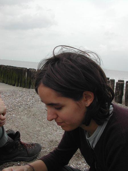 A person with short dark hair looks down while sitting on a beach with wooden barriers in the background.