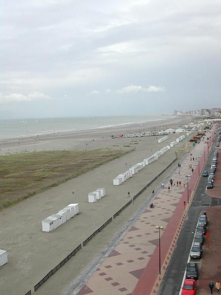 A beachside promenade with people walking, parked cars, and small white beach huts lined up on the sand.