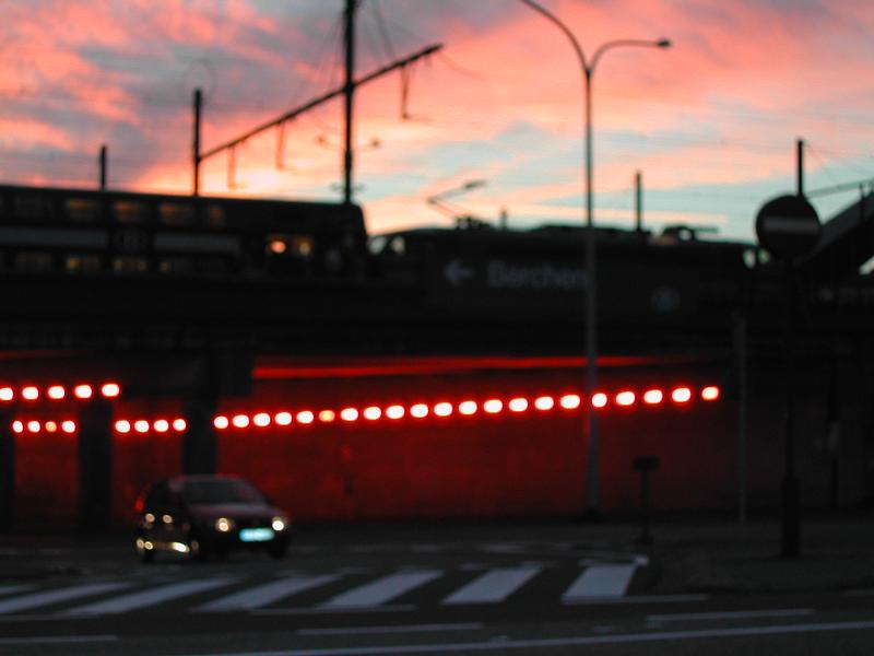 A car drives past a railway bridge at Station Berchem, with red lights illuminating the underpass at dusk.