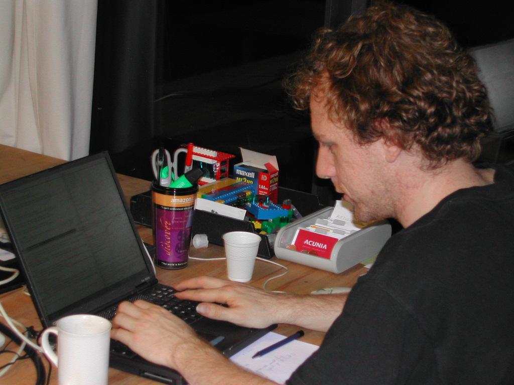 A person with curly hair types on a laptop at a cluttered desk with office supplies and cups.