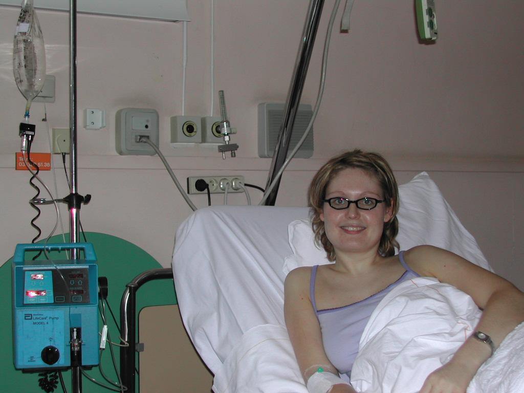 A woman with glasses is lying in a hospital bed, smiling, with an IV drip connected to her arm.