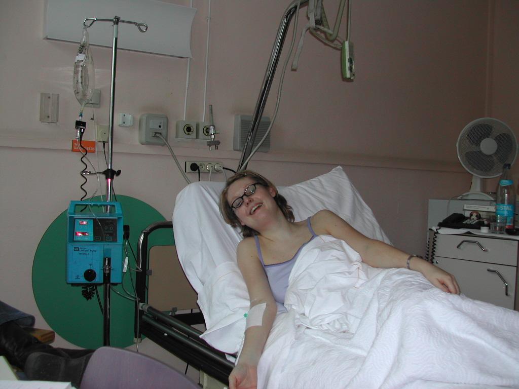 A woman is lying in a hospital bed with an IV drip, smiling and looking towards the camera.