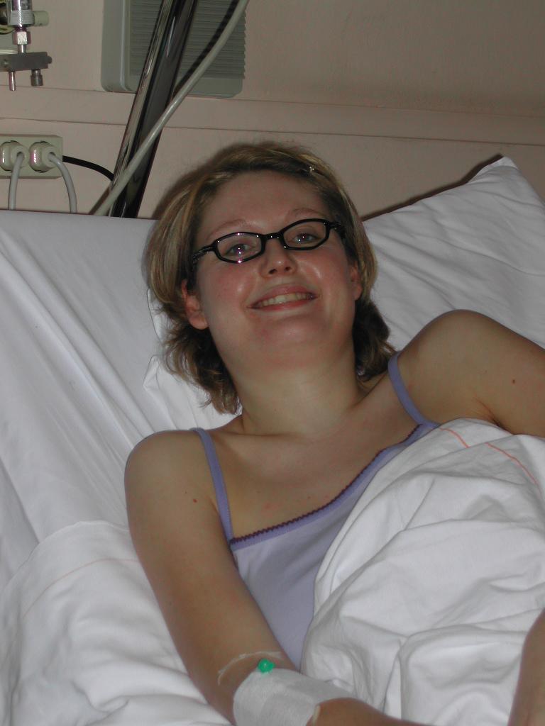 A woman with glasses is lying in a hospital bed, smiling, with an IV in her arm.