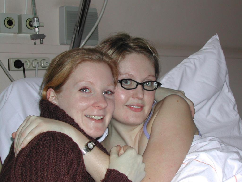 Two women hugging and smiling, one lying in a hospital bed and the other wearing a sweater.