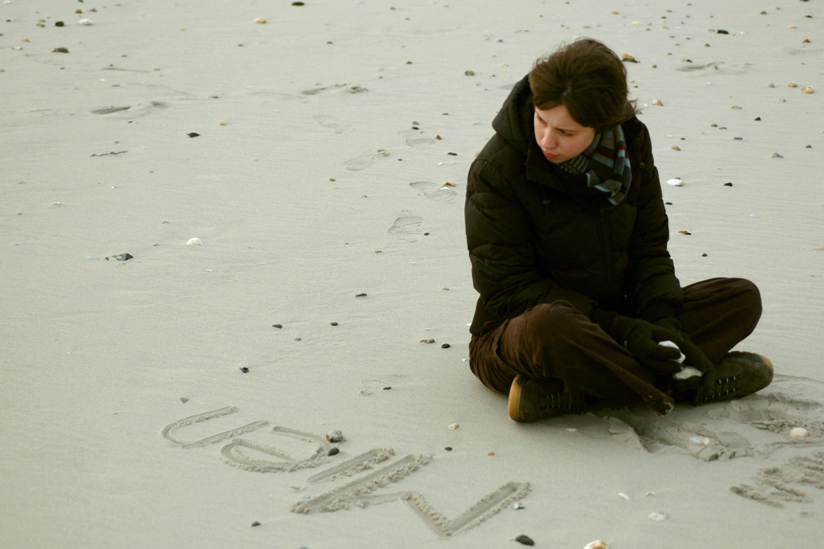 A person in a dark coat sits on a sandy beach, looking at the word "Mien" written in the sand.