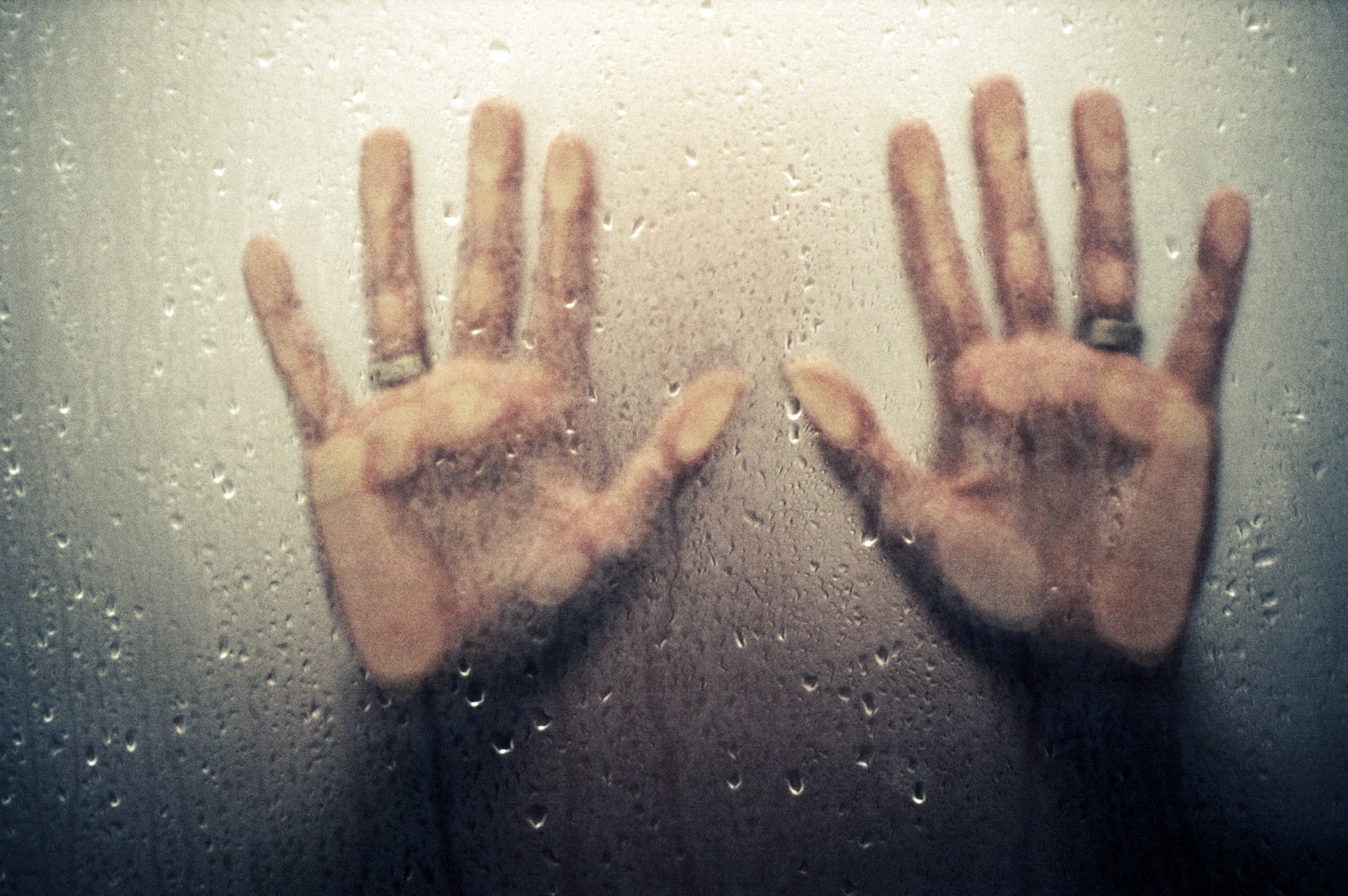 Two hands with fingers spread press against a wet, foggy glass surface, partially obscured by condensation and water droplets.