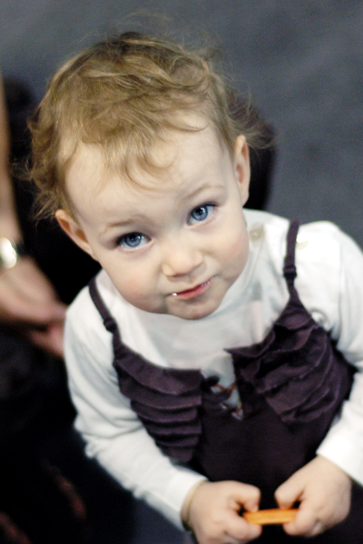 A young child with blue eyes looks up while holding an orange object with both hands.