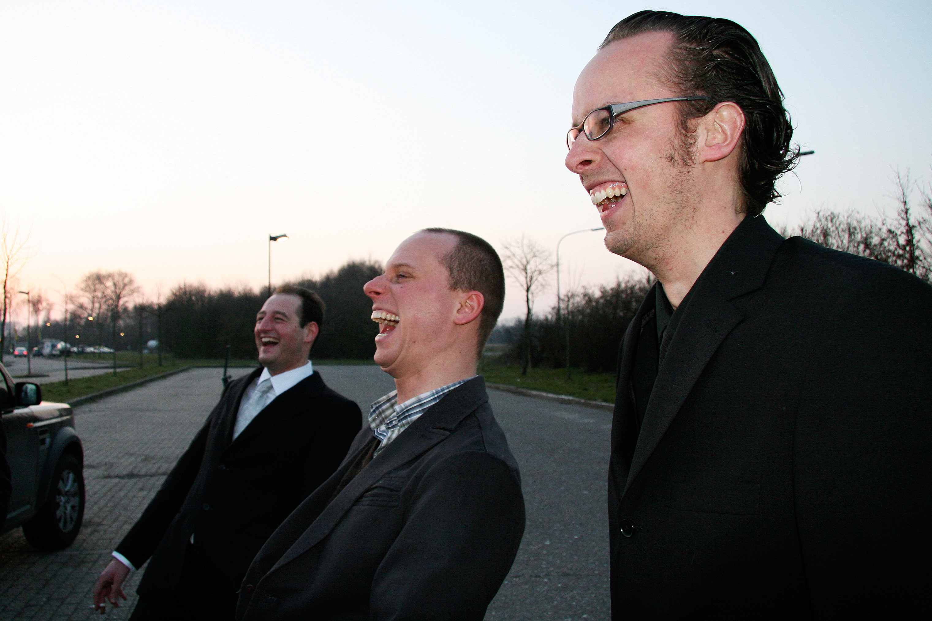 Three men in suits stand outside, laughing together during a bachelor party.