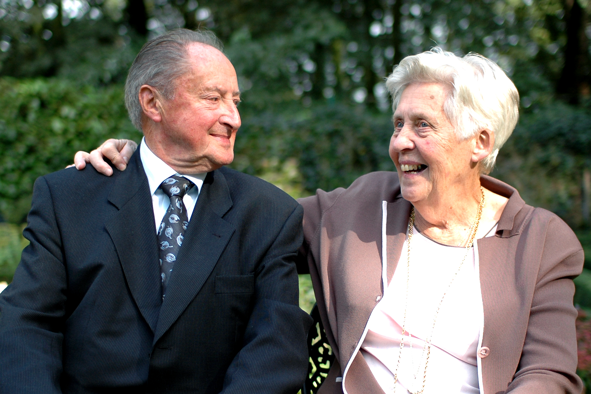 An elderly couple sits together, smiling at each other while celebrating their 55th wedding anniversary outdoors.