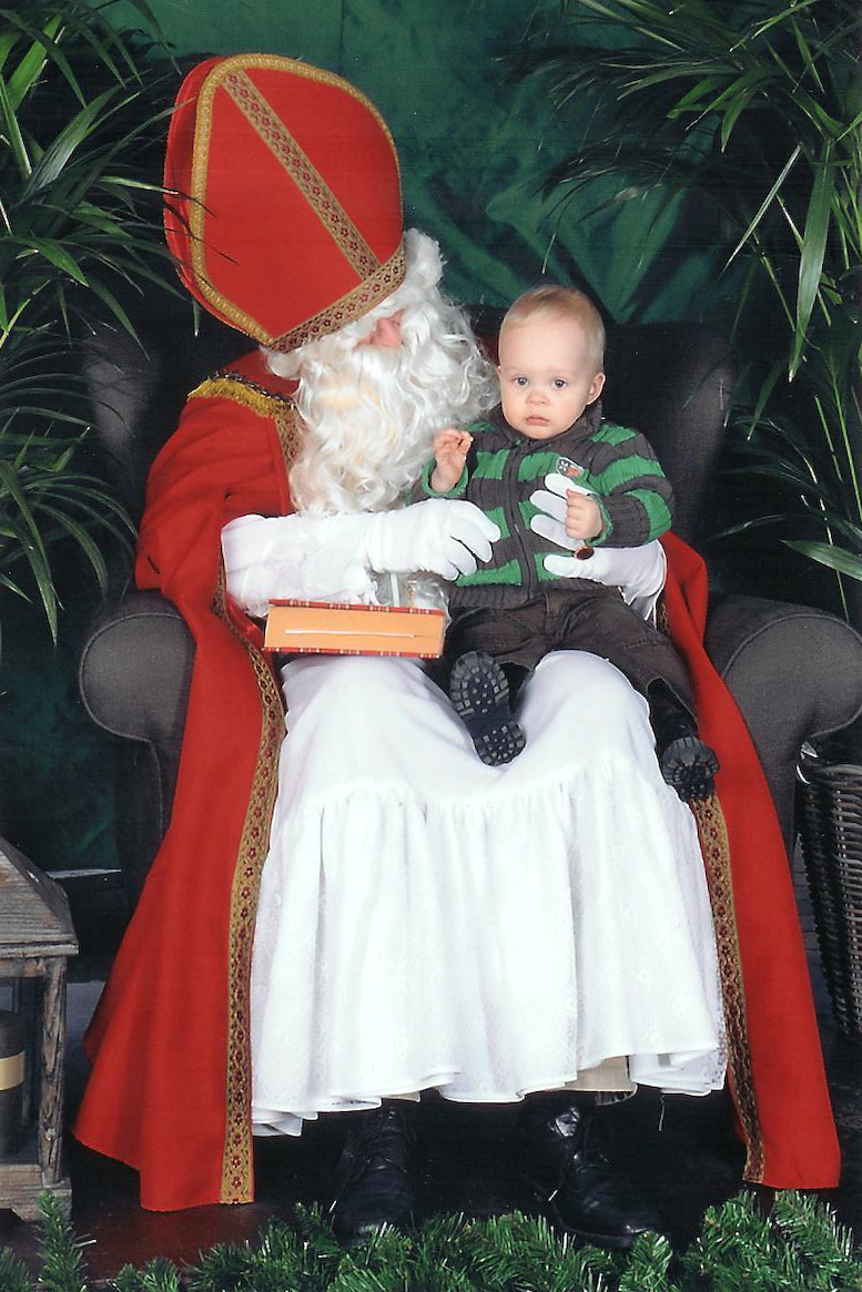 A baby sits on the lap of a man dressed as Sinterklaas, holding a small gift box.