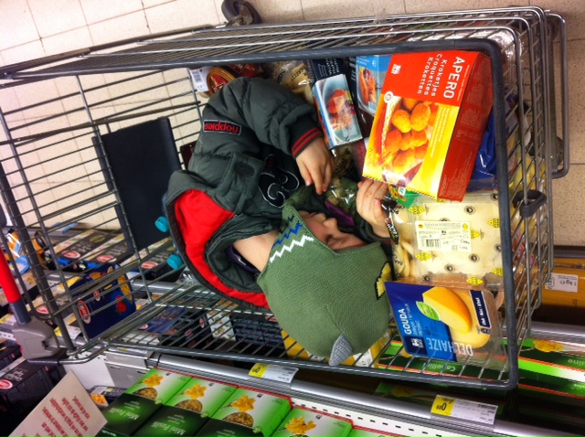 A child wearing a green hat and winter coat is sleeping inside a shopping cart filled with groceries.