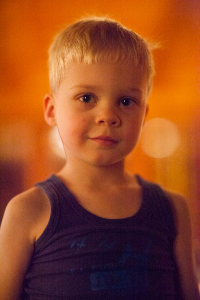 A young child with short blond hair looks directly at the camera, wearing a dark tank top.