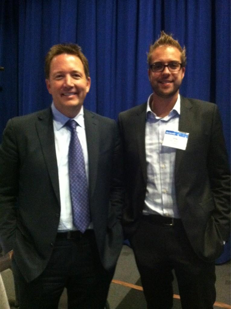 Two men in suits stand side by side, smiling at the camera in front of a blue curtain.