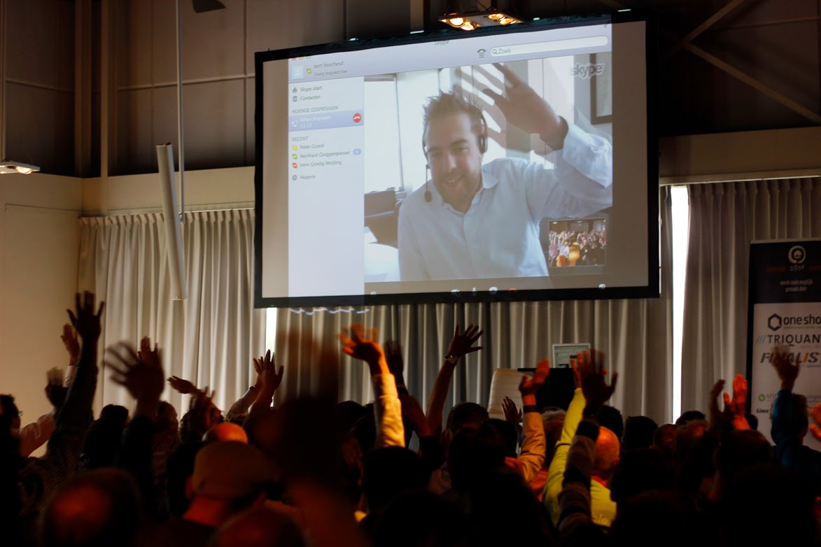 A man on a video call waves on a large screen while a crowd below waves back.