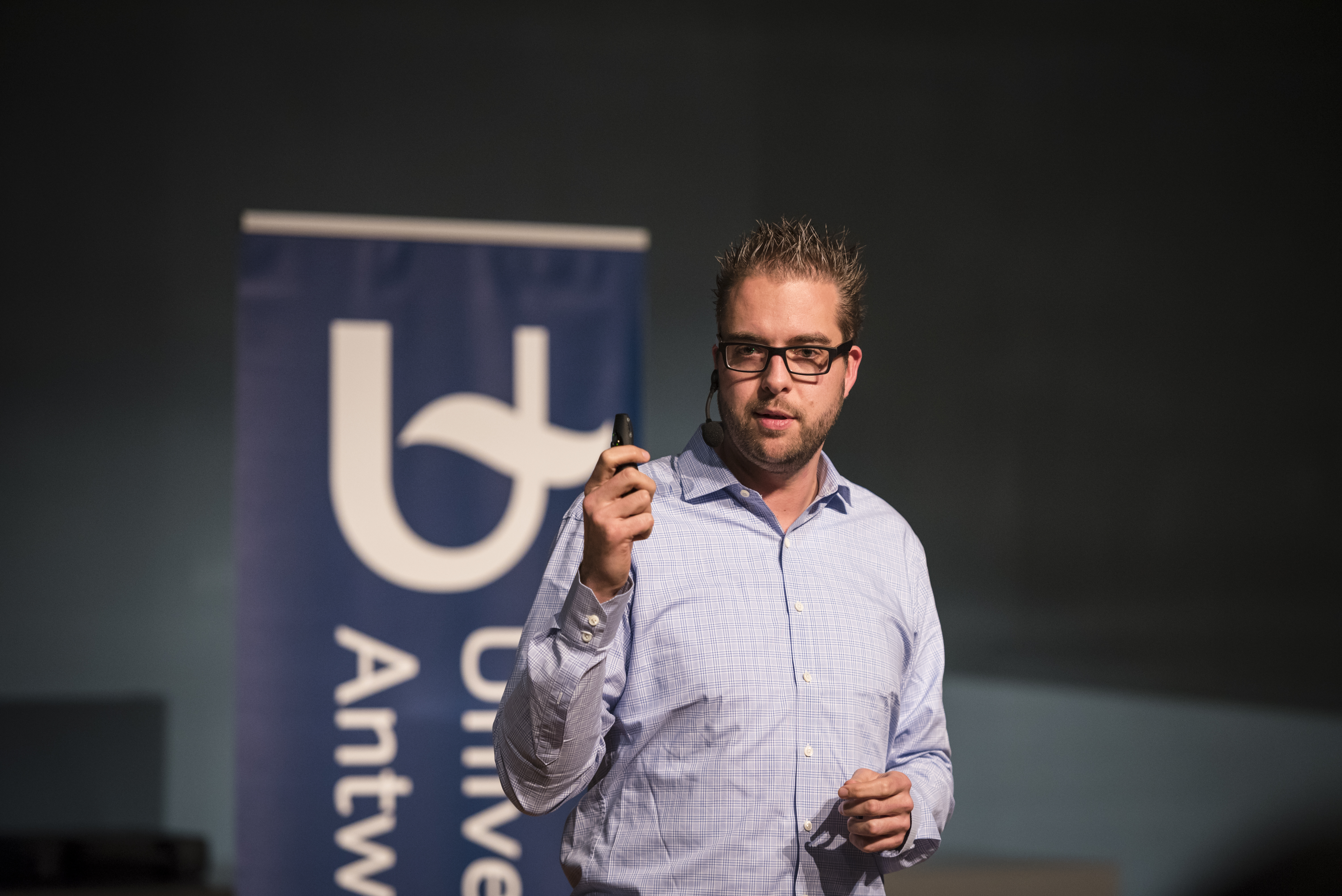 A speaker in a light blue shirt presents at Antwerp University, holding a remote control and wearing a headset microphone.