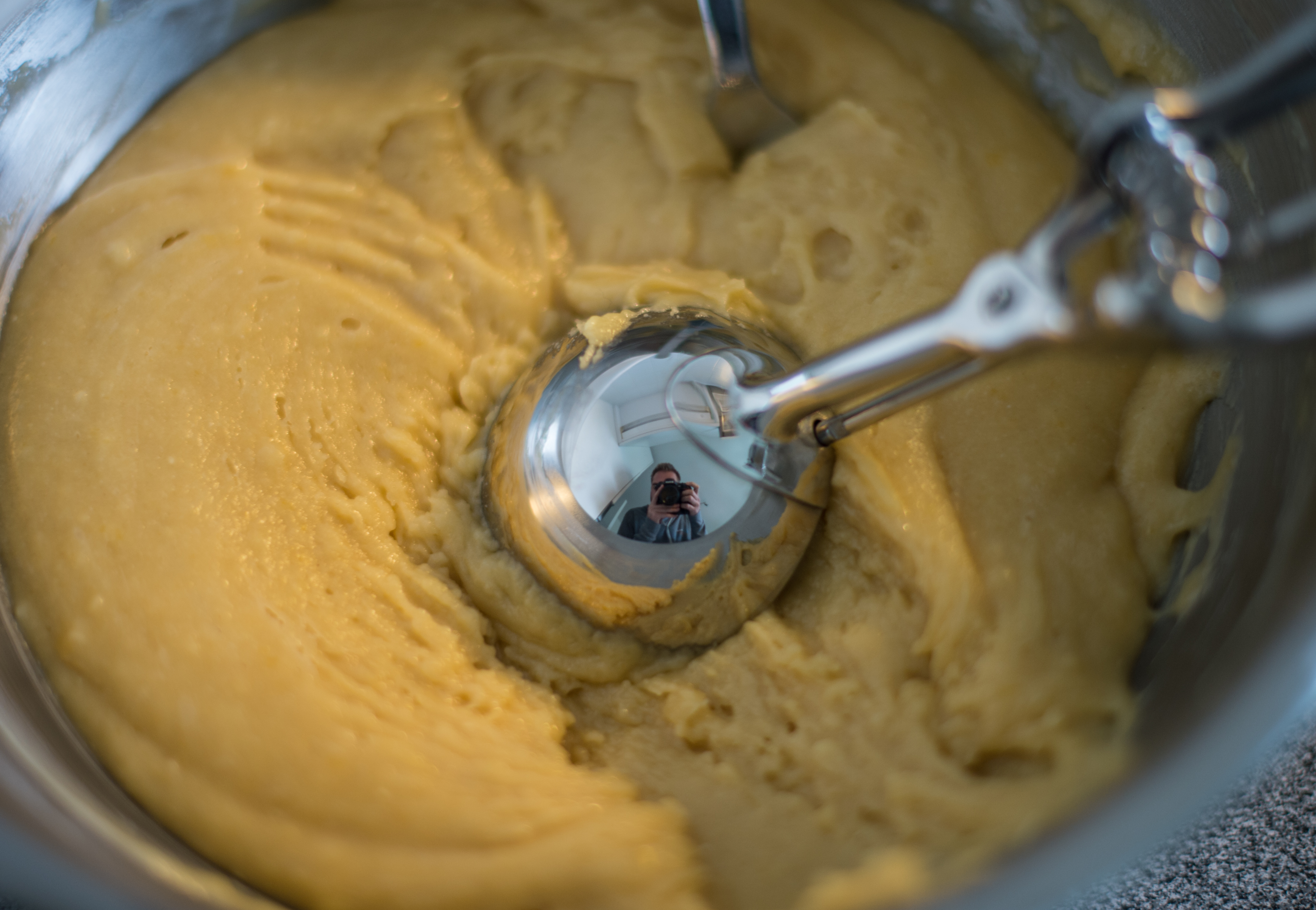 A person takes a selfie reflected in a shiny mixer beater, surrounded by waffle dough in a mixing bowl.