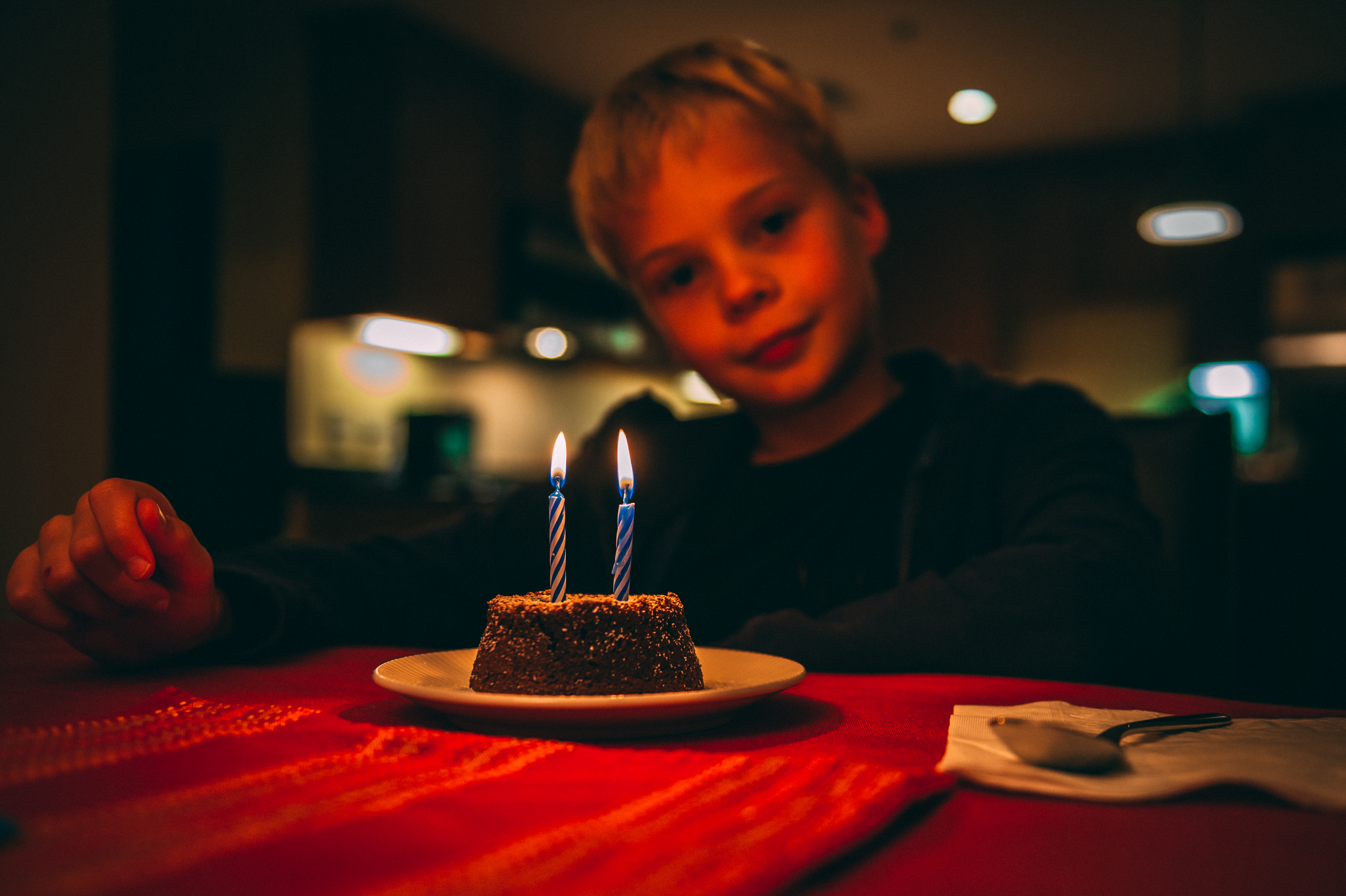 A child sits at a table, looking at a small birthday cake with two lit candles.