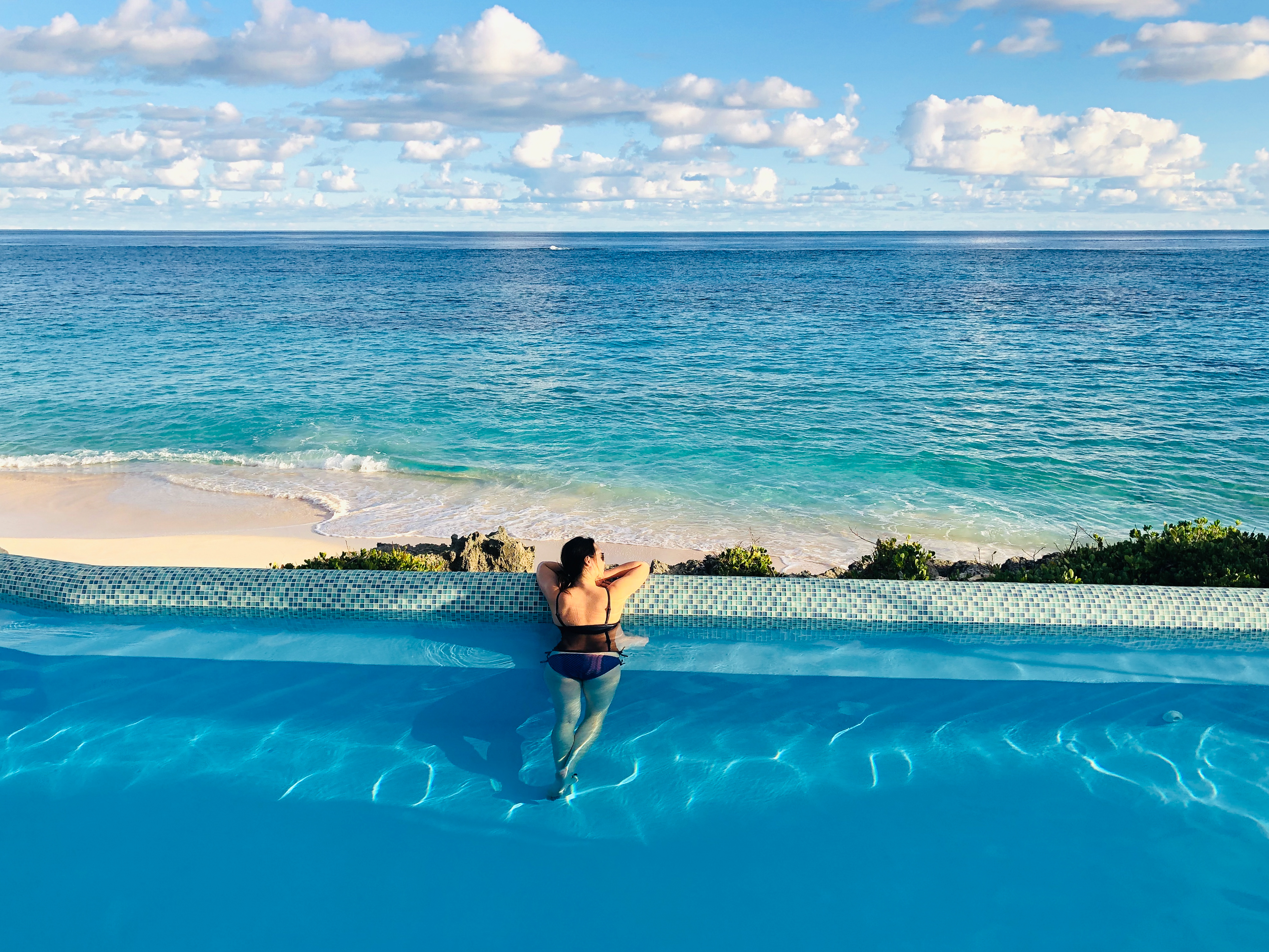 A person in a swimsuit leans on the edge of an infinity pool, looking at the ocean and beach.