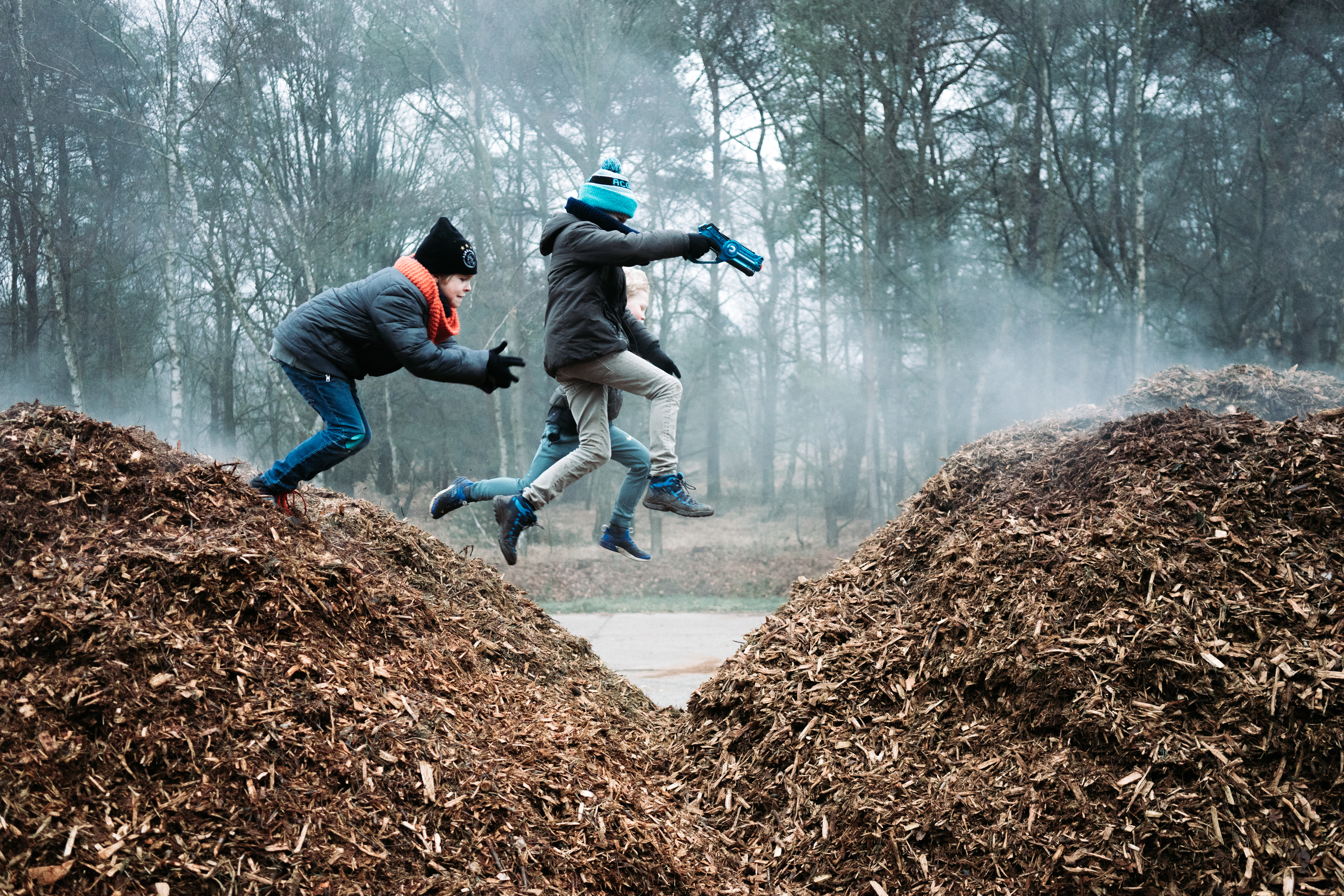 Three children in winter clothing jump between two piles of wood chips, one holding a toy blaster.