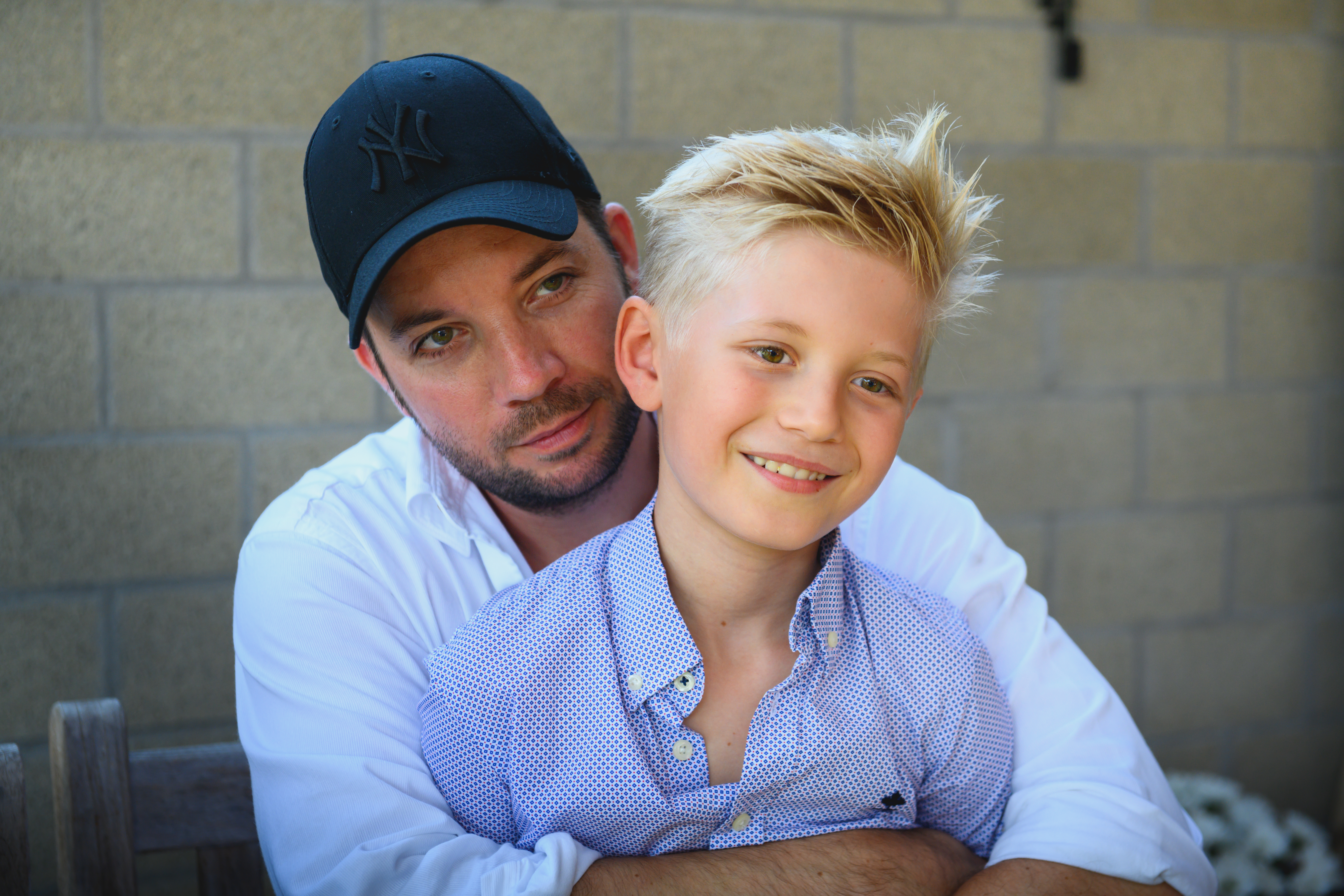 A man in a white shirt and black cap hugs a smiling boy in a blue shirt.